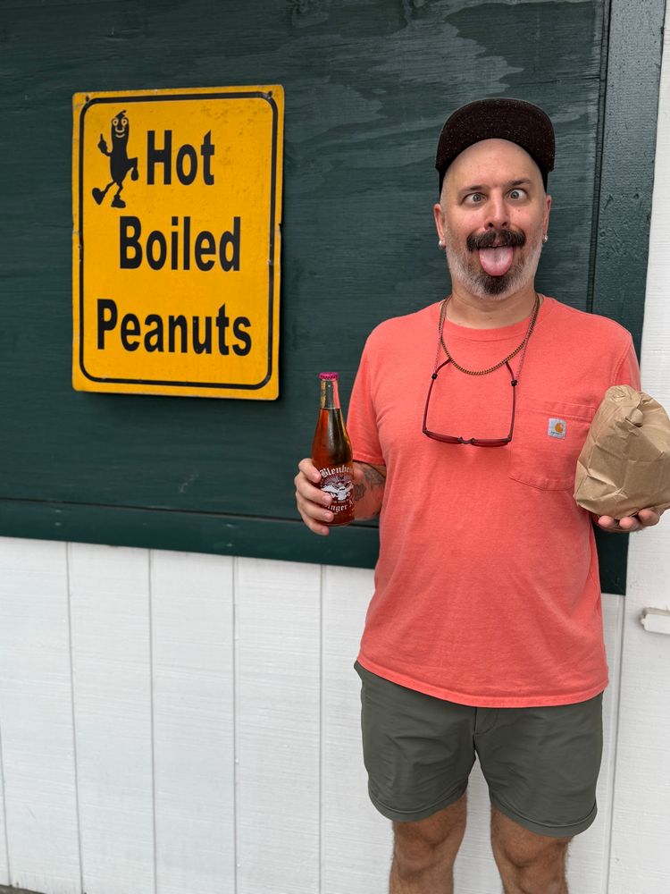 Man with crossed eyes and his tongue out with a bag of boiled peanuts in his hand, posing with a boiled peanuts sign 