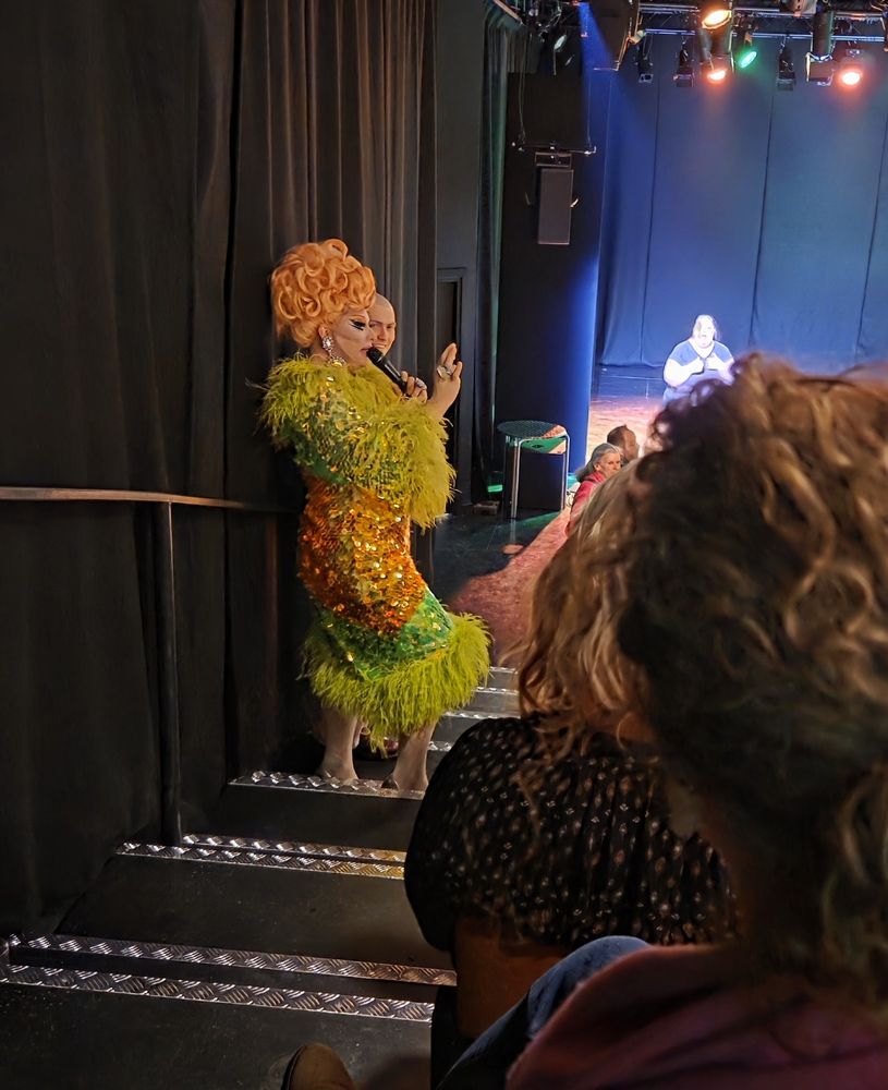 Drag queen Ginger Johnson in a lime green and orange dress, talking into a microphone in a theatre. 