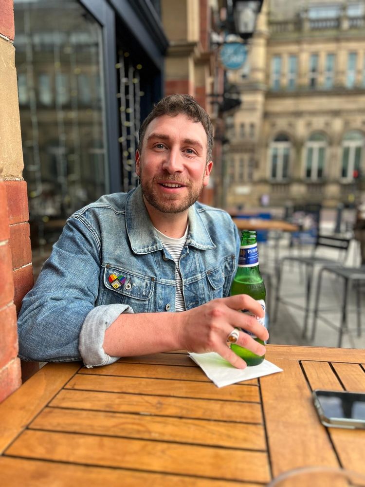 Man in a denim jacket holding a beer smiles at someone behind the camera 