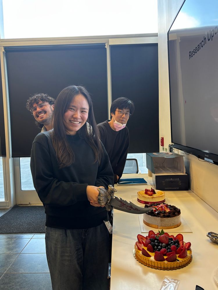 A photo of three people and three cakes. Qian is in the front, holding a serrated knife with dragon accents, poised over a fruit tart. Behind her and bending left is Pal, and behind him and bending right is Skott.