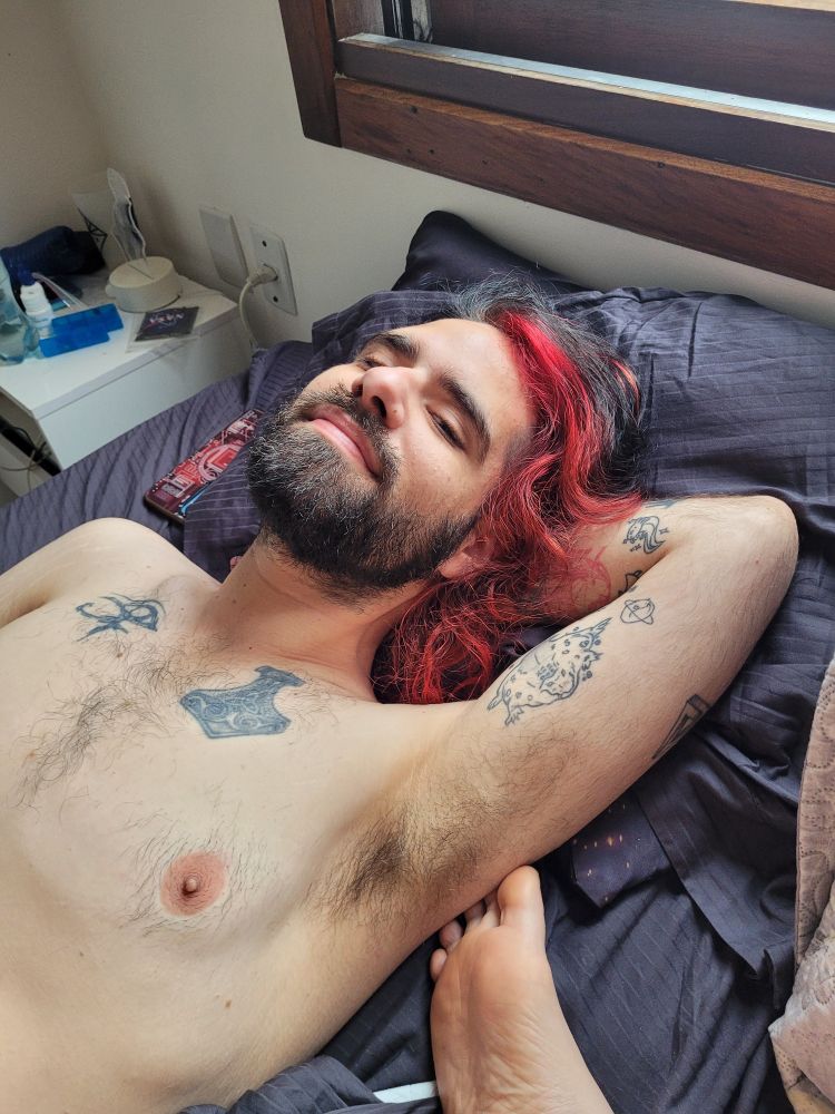 Homem gostoso, deitado na cama sem camisa. Ele tem cabelo comprido, vermelho e preto, barba e sorri para a câmera com os olhos semiserrados.