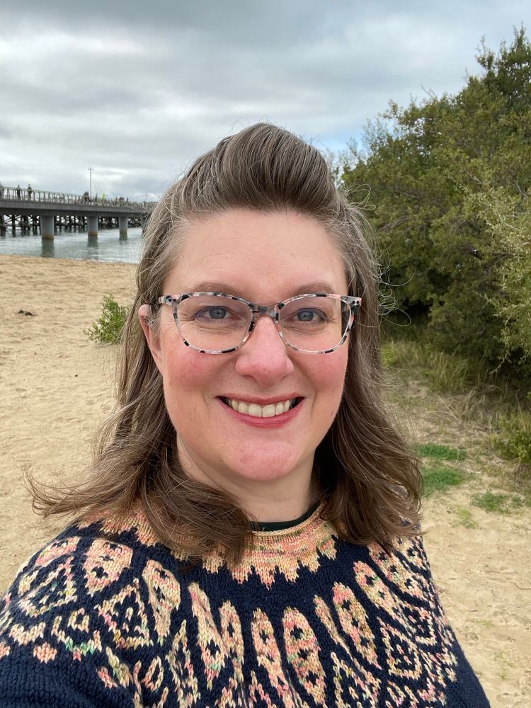 A woman stands in front of a beach and bridge taking a selfie in a navy and peach colour-work jumper.