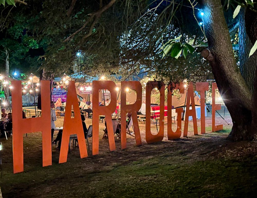 A large orange sign for Harrogate at the Theakston’s Crime Writing Festival, lit up at night, beneath the trees. 