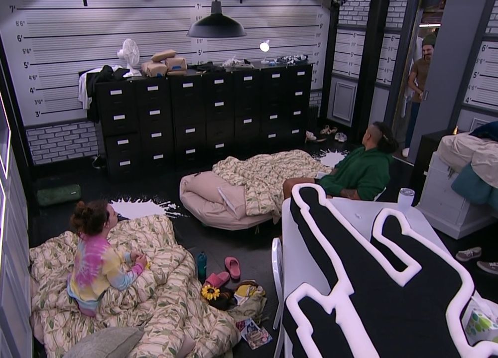 Ava sitting in a bed on a floor, Keanu sitting at a chair behind an outline of a body on a desk, and Vince opening the door to the Have Not bedroom in Big Brother season 27. Vinny is standing in the half open doorway on the outside of the room. The room has mattresses on the floor, a large dresser, and the walls have height markers.