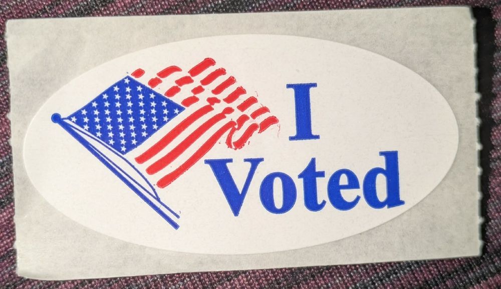 an "I voted" sticker. A white oval sticker on white paper with an American flag and blue words "I voted".