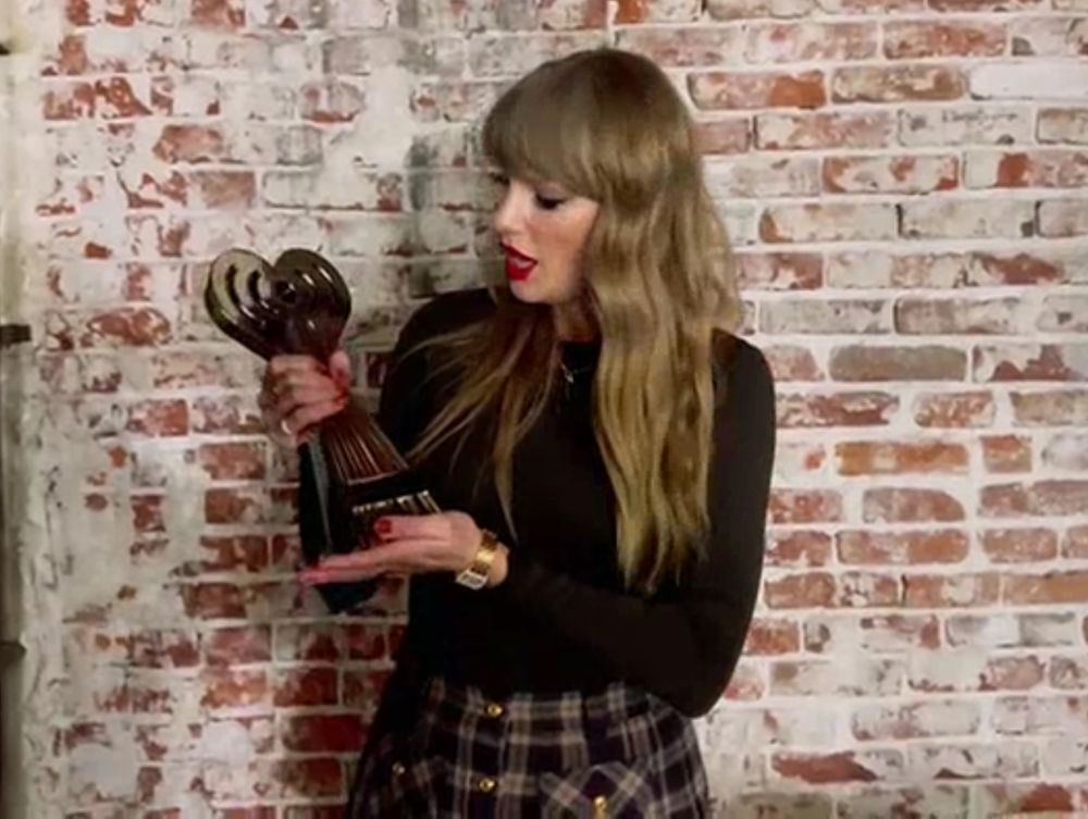 Taylor Swift with a brick wall in the background holding an iHeartRadio music award. She's wearing a black shirt and a brown plaid skirt. 