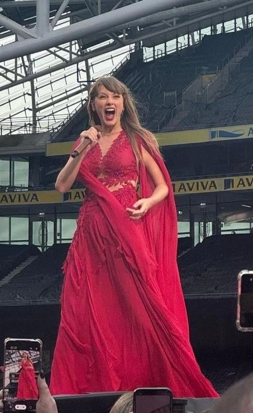 Taylor Swift with a huge smile on her face while performing on the Eras Tour because she spotted Travis Kelce arriving. She's wearing a red folklore dress and holding a microphone.