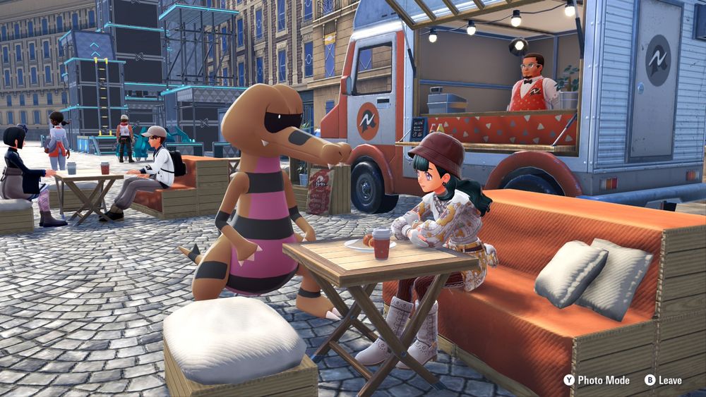 Pokémon Legends Z-A game screenshot. My character is sitting on an orange bench at an outdoor food truck cafe with a Krokorok standing next to her. There's drinks and food on the table in front of my character and people and buildings in the background.