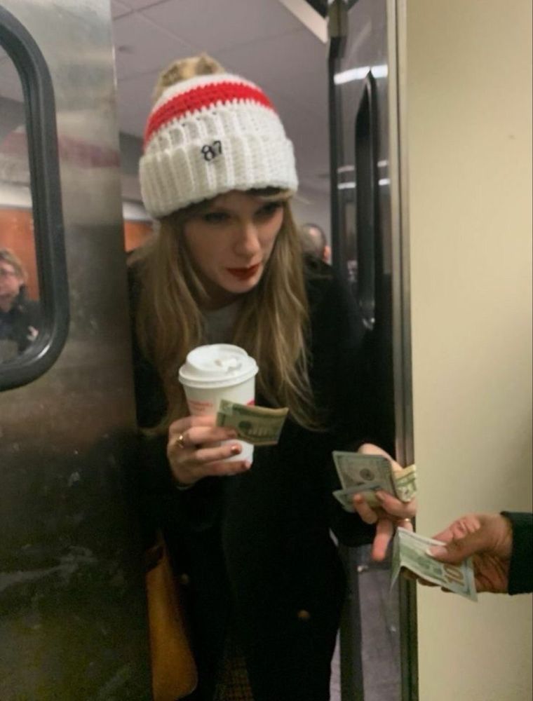 Taylor Swift in a winter hat is handing someone cash through a half open door while holding coffee and more cash in both of her hands.
