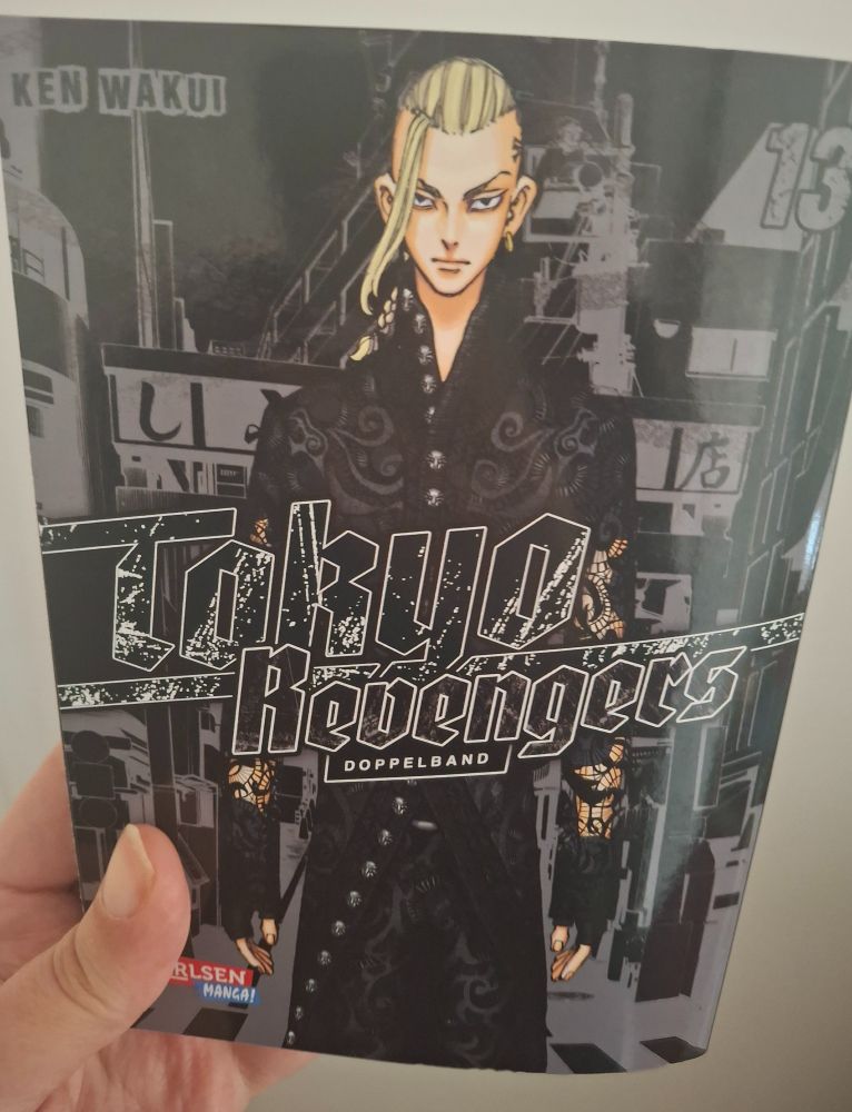 a left hand holding a manga; on the cover is a tall young man, blond hair, black clothing and seriously looking. The title says: Tokyo Revenger (two volumes).