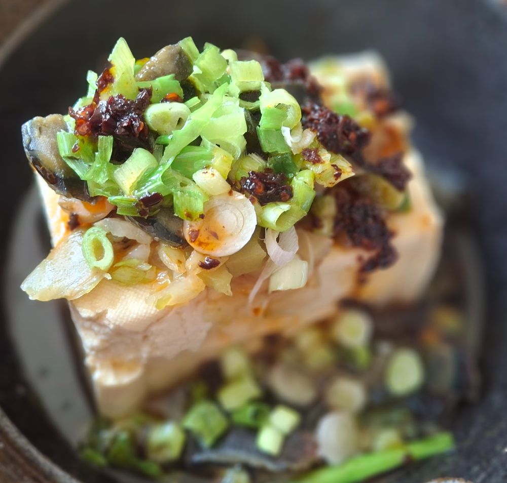 a black ceramic bowl with a block of cotton tofu, topped with chili oil, a chopped century egg and scallions.