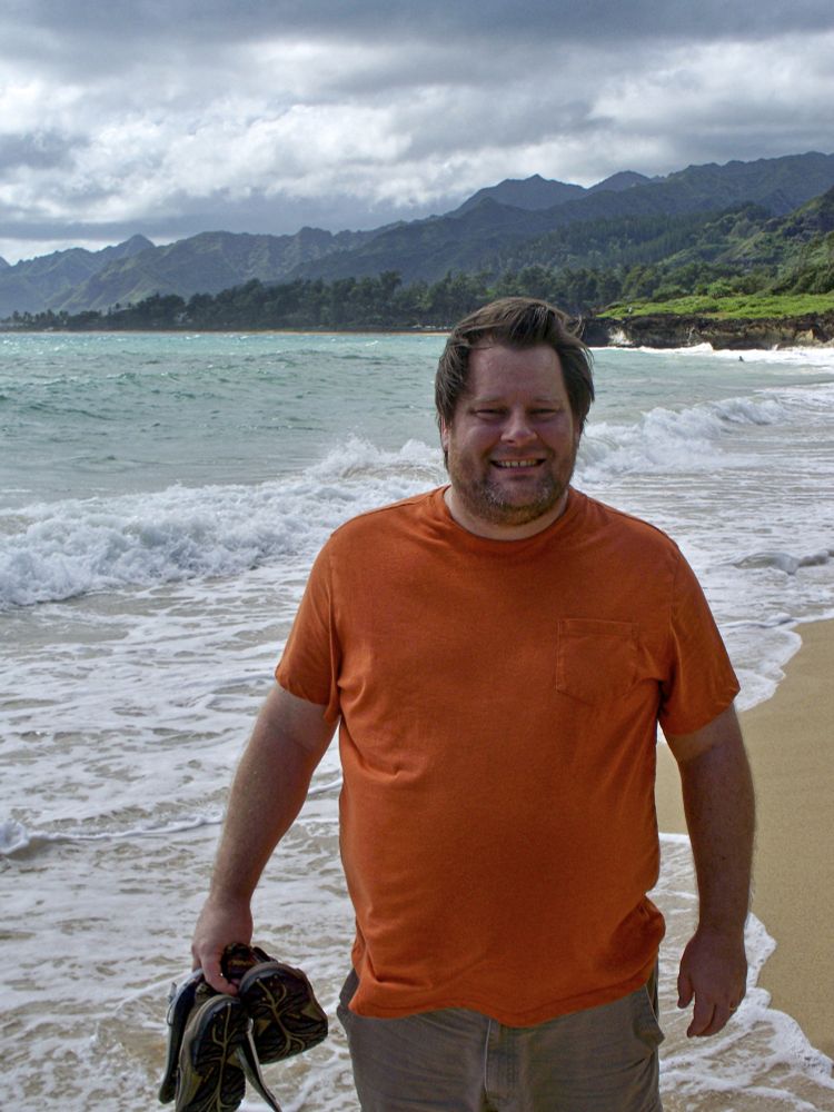 A large bearded man in an orange shirt stands in front of a beach in Hawaii.  He is clearly overweight and in poor health.