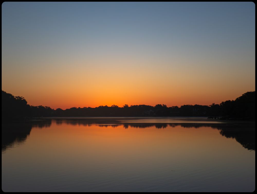 Photo shows a calm still lake with a dark treeline covering the entire opposite coast, with taller trees towards the edges of the frame on each side. Almost looks symmetrical.  The sun has not quite risen but the colors of it coming fill the frame, with oranges along the treeline giving into light yellows and blues further up.  All is reflected in the lake very nicely.
