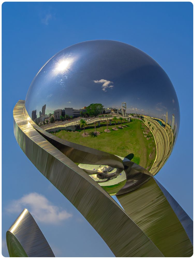 A sculpture that appears to be made of steel.  It is a perfectly round globe held up by twisting arms that come from the bottom right of the frame.  Behind the sculpture is clear blue skies and reflected in the sculpture is a mini-city scene with interstate on the right and bulidings and a pedestrian bridge on the left with a small green area between.