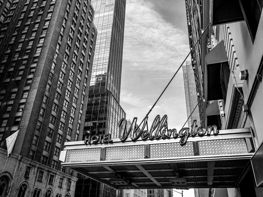 A black and white photo of the "Hotel Wellington" marquee hanging at the bottom of the frame.  It is surrounded by tall buildings on either side and the gray sky peeks through the center background.