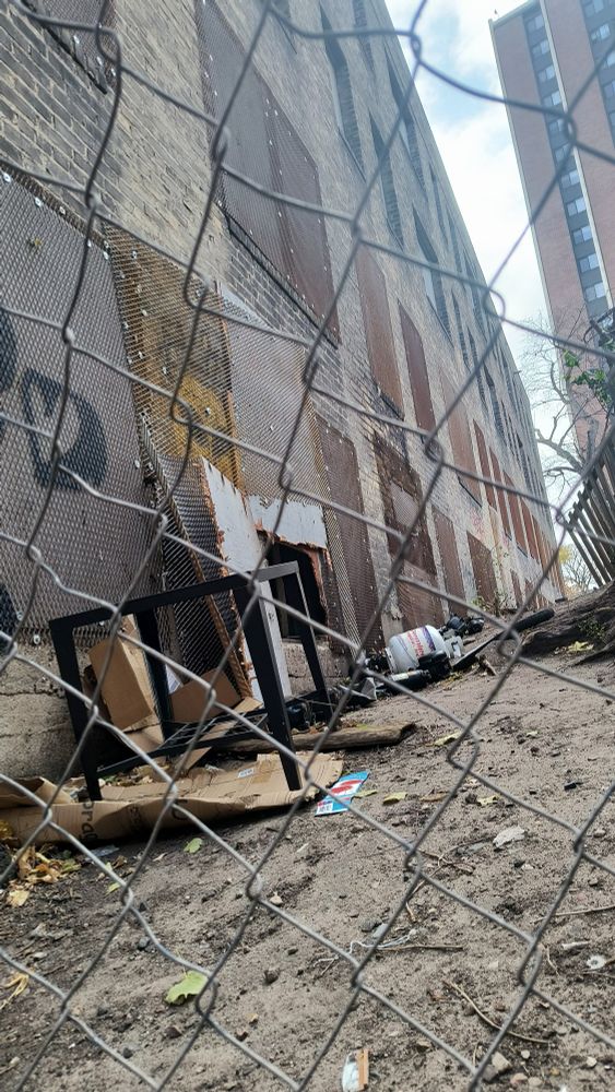 Abandoned building with steel grates over window and propane gas tank next to hole cut thru cage and board over window