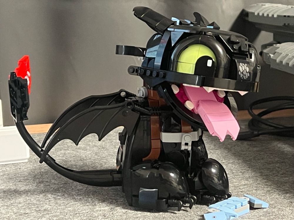 Lego version of the Nightfury dragon toothless sitting upright, open mouth, tongue out and fish in front of him.
His head is turned slightly to the left