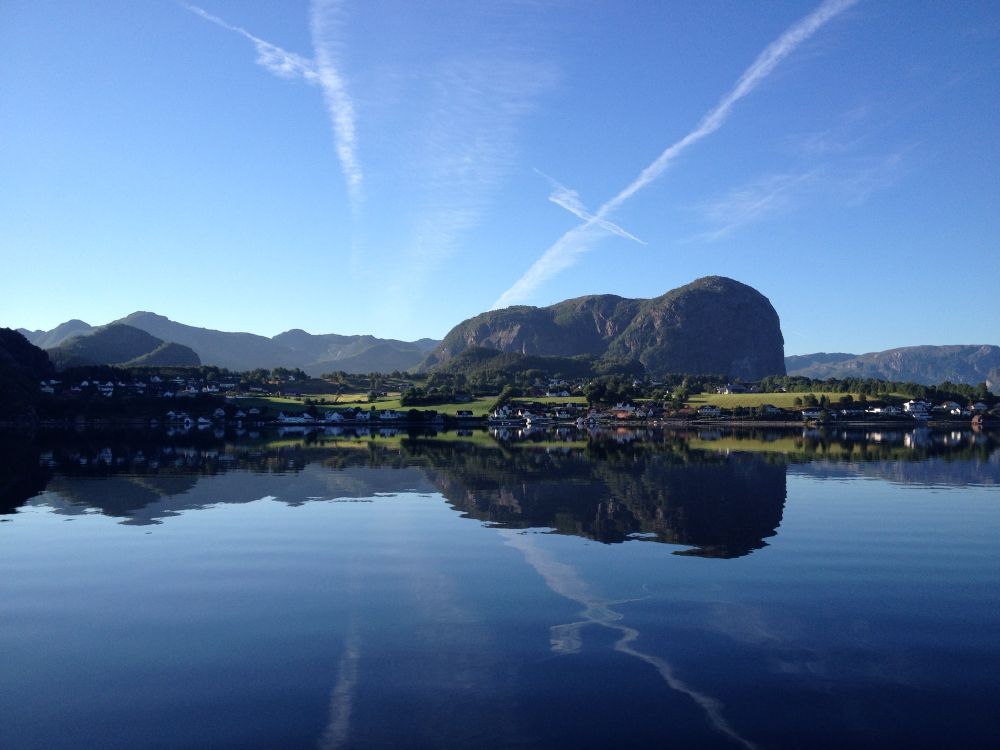 Blick von einem Schiff in Richtung Land. Das Wasser ist spiegelglatt und reflektiert das Ufer und den Himmel fast komplett. Selbst die weißen Kondensstreifen am hellblauen Morgenhimmel reflektieren sich ich dunkleren Blau des tiefen norwegischen Fjord 