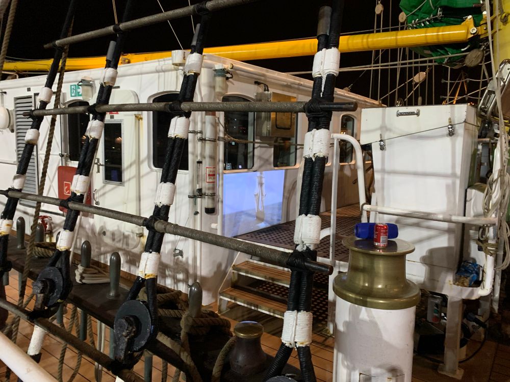 View from just outside a tallship onto the deck. Looking through the shroudes onto map and navigation housing. On the white wall you can see a movie being projected, an shot a classic tall ship sailing away from the camera. It’s the HMS surprise from Master and Commander