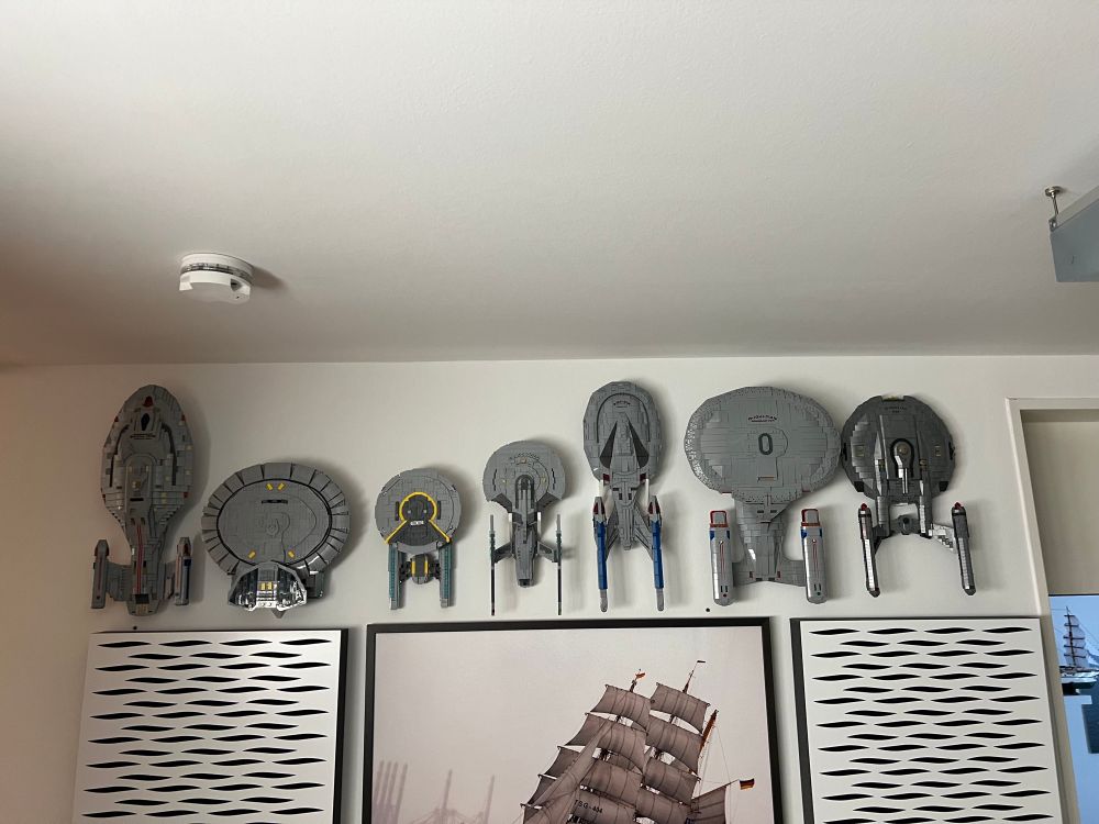 7 brick build Lego compatible spaceships, lined up and wall mounted, from left to right: USS Voyager by BlueBrixx, a Nebular Class, USS Ceritos, USS Enterprise G all by lilium brick yard, USS Enterprise E, D and NX-01 BlueBrixx