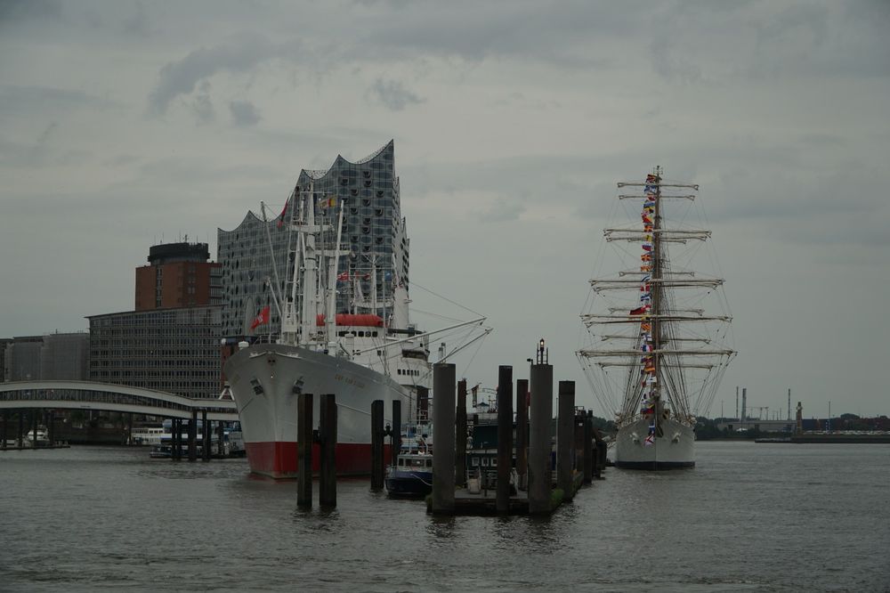 The Argentinian tallship A.R.A. Libertad in Hamburg. The white ship with its tall masts on one side of the Überseebrücke (Overseabridge) with the so called white swan of the south Atlantic on the other side of the pier. The Cap San Diego a still sea worthy museum ship. In the background, the iconic Elbphilharmoni. The sky is in a typical joyful summer grey.