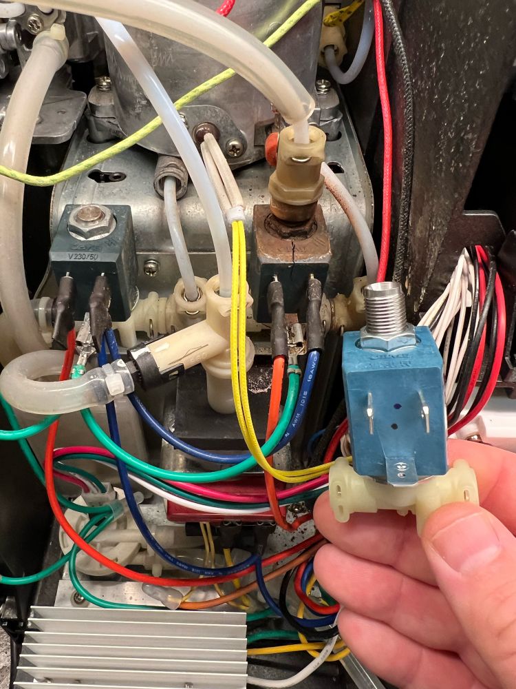 View of the inside of a coffee machine with a chaotic but colorful wiring. In between a lot of tubes for water and steam. One valve is noticeably dirty and has a cracked housing. In comparison I’m holding a fresh new valve right next to it
