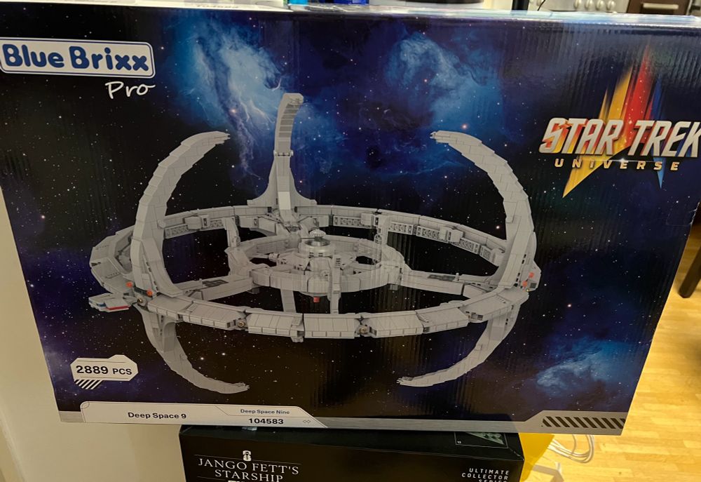 A sealed box of a brick build Star Trek Deep Space 9 Set with 2900 pcs.
The package is held und black and dark blue background and the cardassian fascist eyesore right and center. A small Defiant is docked on the docking ring.
Unfortunately this set does come with neither Jadzia nor Ezri Dax