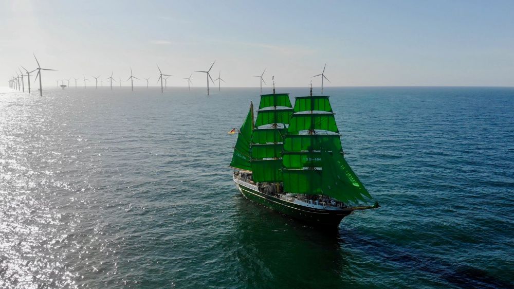 Der Großsegler Alexander von Humboldt 2 mit seinen markanten grünen Segeln vor einem Offshore Windpark bestehend aus Dutzenden Windkraftanlagen die ordentlich aufgereiht über das Meer erstrecken 