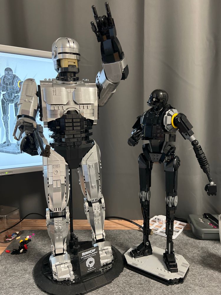 Brick build (Lego) K-2SO from Star Wars standing next to brick build (BlueBrixx) Robocop. K-2SO is looking upwards to the significantly larger Robocop how holds his weapon in the right hand and has his left arm raised high giving metal sign
