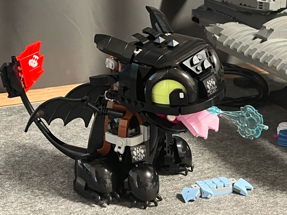 Lego version of the Nightfury dragon toothless sitting upright, open mouth with a blue flamed breath and fish in front of him