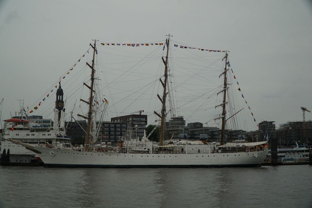 The Argentinian tallship A.R.A. Libertad in Hamburg. The white ship with its tall masts watched from the water side. In the background one of Hamburgs famous church towers can be seen the „Michel“. The sky is in a typical joyful summer grey.