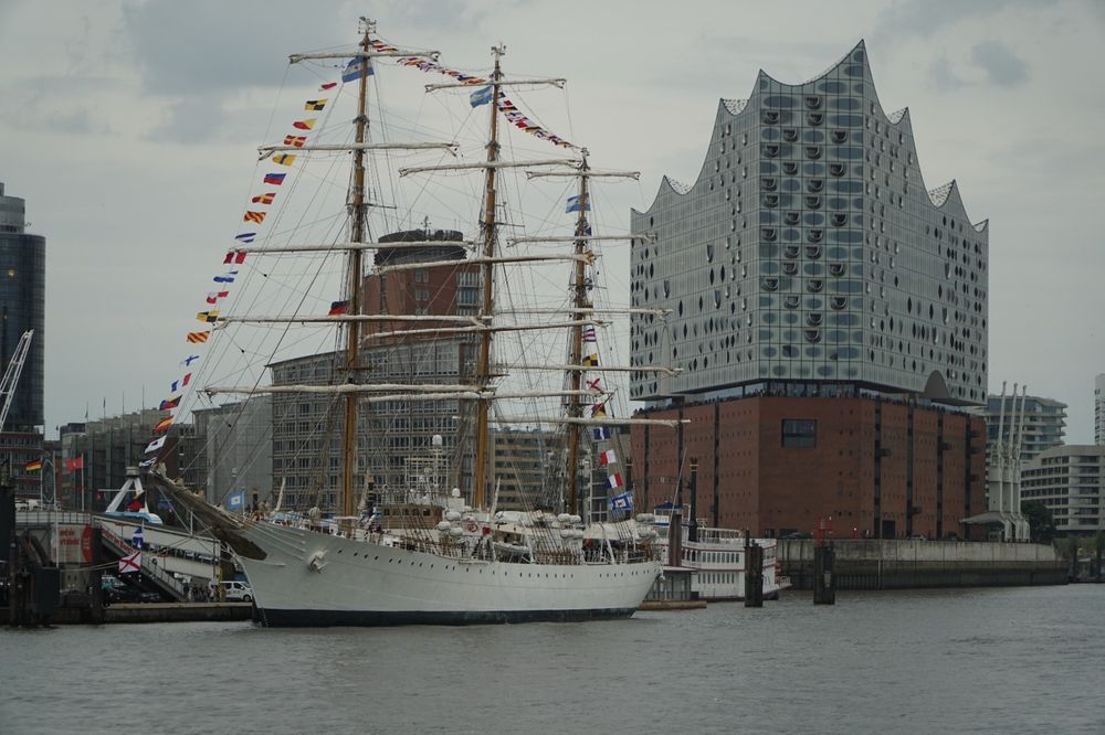 The Argentinian tallship A.R.A. Libertad in Hamburg. The white ship with its tall masts watched from the water side in front of Hamburg gals-palace-of-music the Elbphilharmoni. The sky is in a typical joyful summer grey.