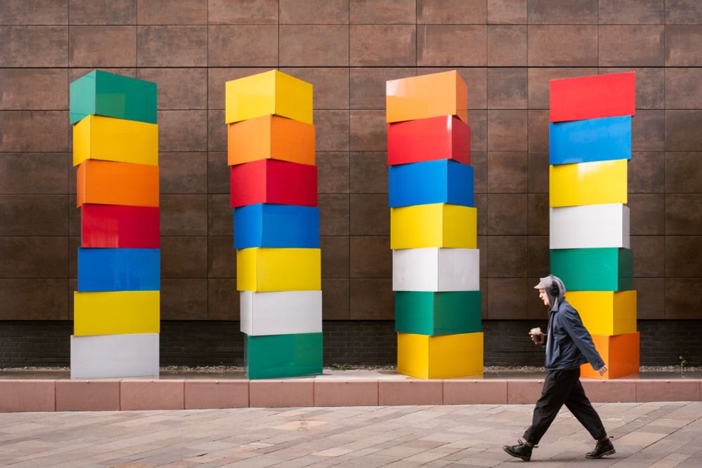 Salford, England. Photograph of part of a sculptural art installation on Greengate titled 'Slow Revolution' by Liz West. The work features a number of stacks of brightly coloured boxes turned so that the edges overlap giving the appearance of twisting or of them being in motion. To the right of frame in the foreground a person is walking by. 