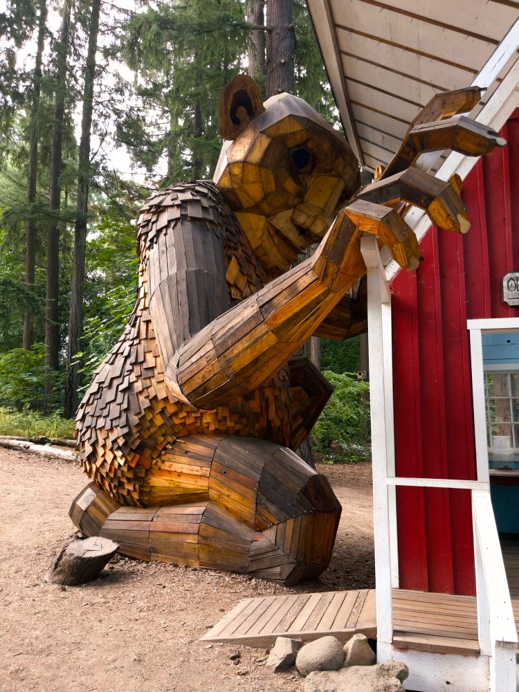 Wooden architectural sculpture of a troll larger than a small house that it is looking into from above through a turned up roof half.