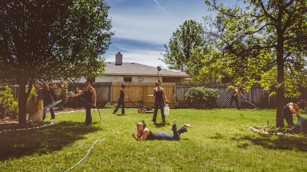 Several versions of the same woman (as if she has cloned herself) are doing various yard work tasks (watering, mowing, etc.) in a suburban backyard. One of them is just lying in the middle of the lawn, playing with dandelions.