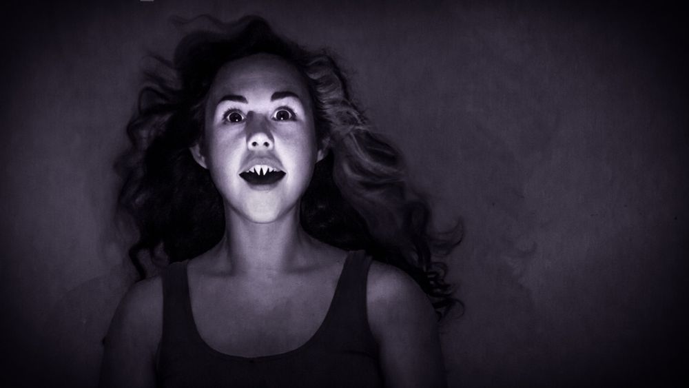 A monochrome image of a woman with very large eyes and teeth that look like fangs - long and ending in points. With eyes wide and eyebrows raised, long hair almost floating up around and behind her, her mouth is ajar in an open mouthed smile, and she is staring straight ahead at you with *great interest.*