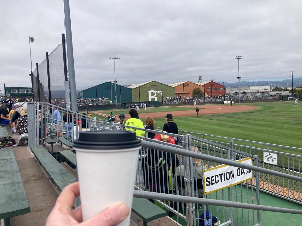 A hand holds up a covered paper cup in front of an Oakland Ballers baseball game on a cloudy afternoon at Raimondi Park. 