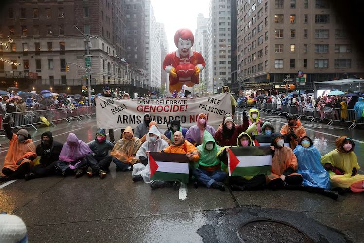 The photo shows a group of protestors sitting in the middle of a wet street during what appears to be a large parade. The group is holding Palestinian flags and a large banner. The banner reads: 