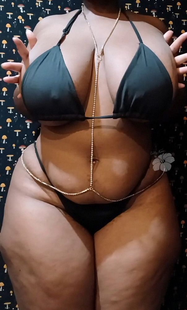 Mauve stands in front of a black background with mushrooms printed over it wearing a black bikini and gold body chain. She pushed her breasts together. Her finger fan out at either side of her breasts. 

#bbw #alt #NsfwSky #SpicySky #goth 