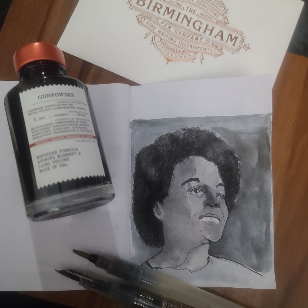 A black-and-white ink portrait of a person (Nikki Giovanni) with curly hair and a smile is drawn on an open sketchbook. Beside the sketchbook is a bottle of 'Gunpowder' ink from Birmingham Pen Company, along with two water brush pens. A branded envelope from Birmingham Pen Company is partially visible in the background.

