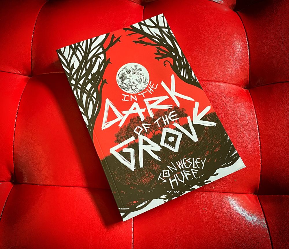 In the Dark of the Grove by Jon Wesley Huff. The cover of the book depicts a red sky with a moon over a cropse of trees isolated in a field. Graphic tendrils sweep upward. The book sits on a red viny cushioned packing.