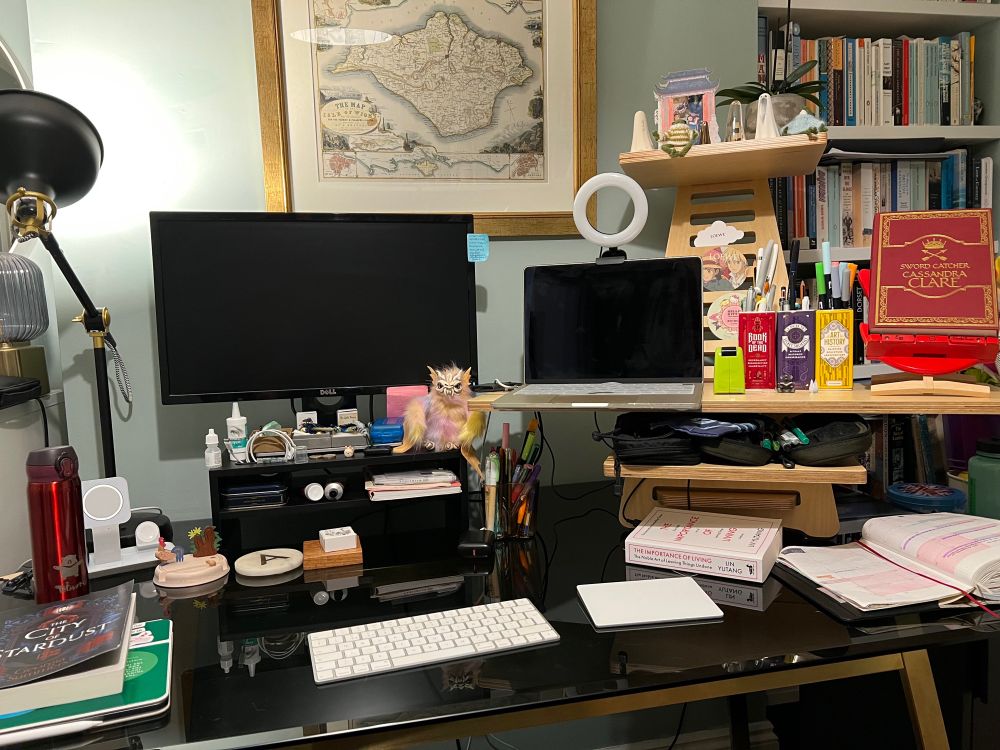 A dark glass desk with a monitor and laptop on a standing desk with Cassandra Clare’s Swordcatcher on a bookchair and lots of tchotchkes like small ceramic ghosts and a skelefloof creature with books and a red thermos and a framed map on the wall.