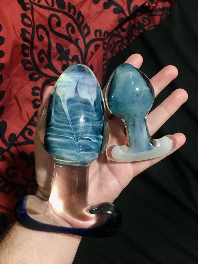 two blue glass anal plugs held in the hand. one is much larger than the other with a circumference of roughly 7", the smaller plug has a circumference of 5"