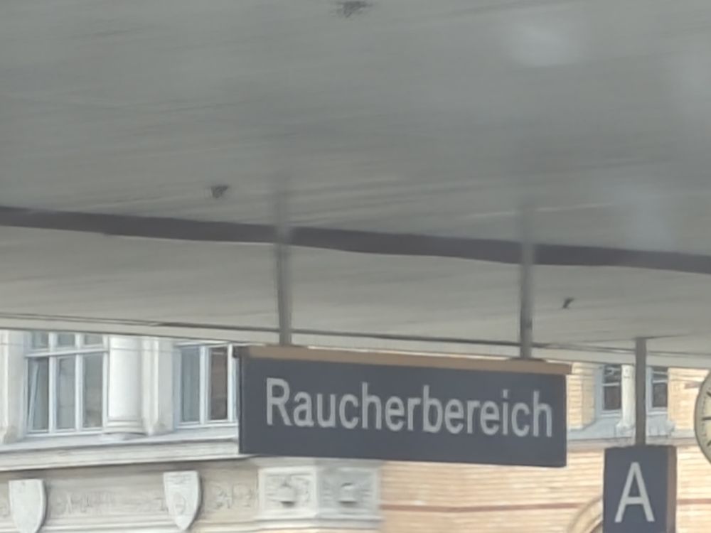 Ein Schild an einem Bahnhof. Es ist genauso groß und in den gleichen Farben gehalten wir die Schilder die anzeigen, in welchem Ort eins sich befindet. Auf dem Schild steht "Raucherbereich".