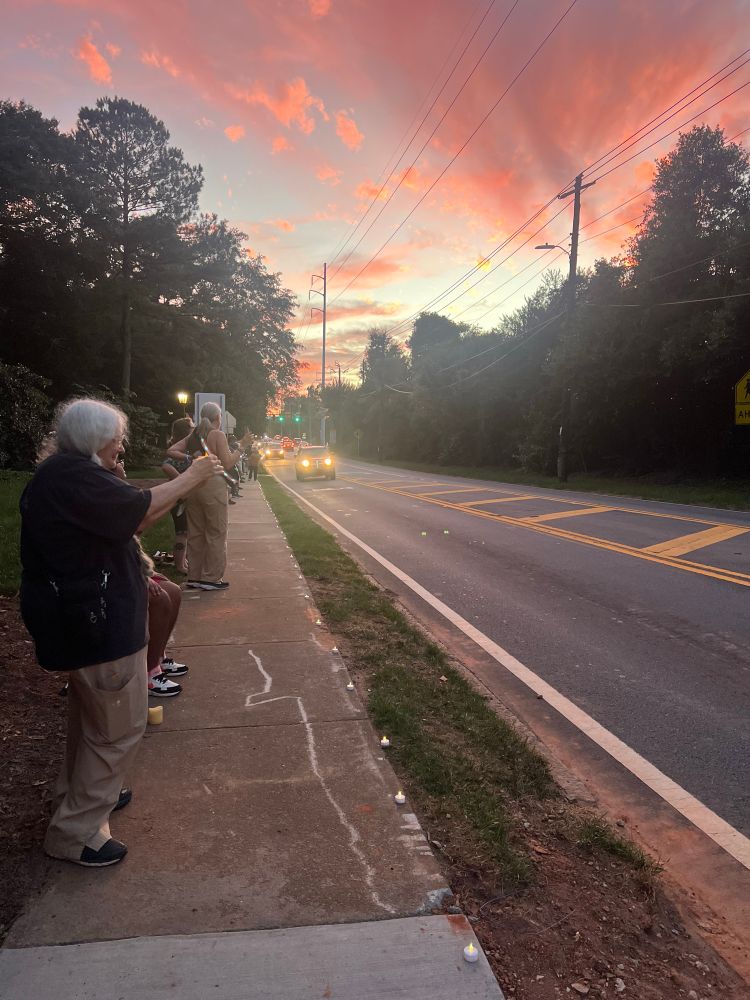 Street view with sunset in the background and people lining the sidewalk with candles and other lights; little LED lights”candles” are spaced along the sidewalk.