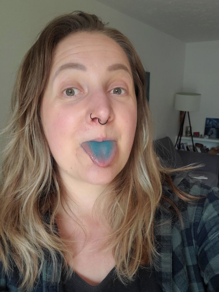 Ash, a white person in their 30s with chest length blonde hair. They're wearing a green flannel shirt. Their tongue is sticking out and it's very blue