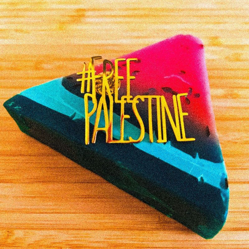 木目の机の上に、スイカの色形をした三角形の石鹸が置かれている。石鹸の上には、金色の「#FREE PALESTINE」の形をしたブローチがある。
