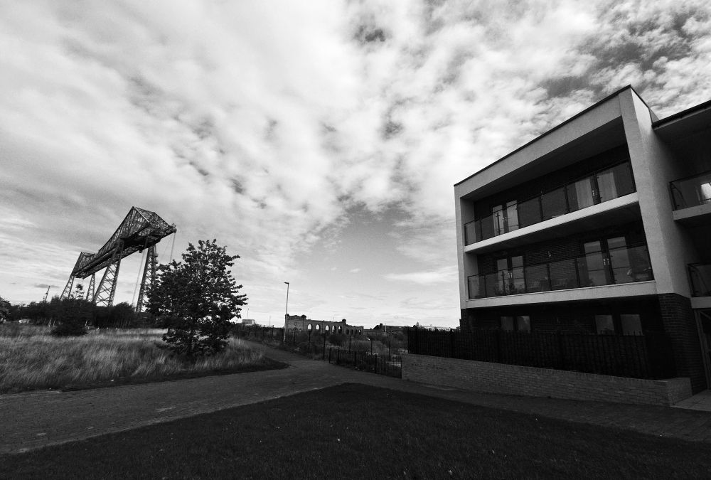 A black and white picture of the Middlesbrough Transporter bridge, a new build and the ruined wall of the old salt works. A wide angle with the clouds moving towards the perspective point
