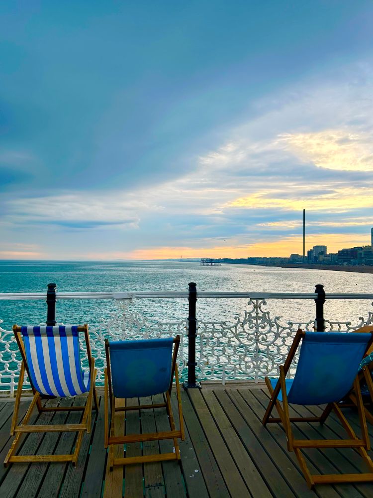 Zwei blaue und ein blau-weissgestreiftet leere Liegestühle auf einem Pier, von hinten fotografiert mit Blick aufs Wasset.
