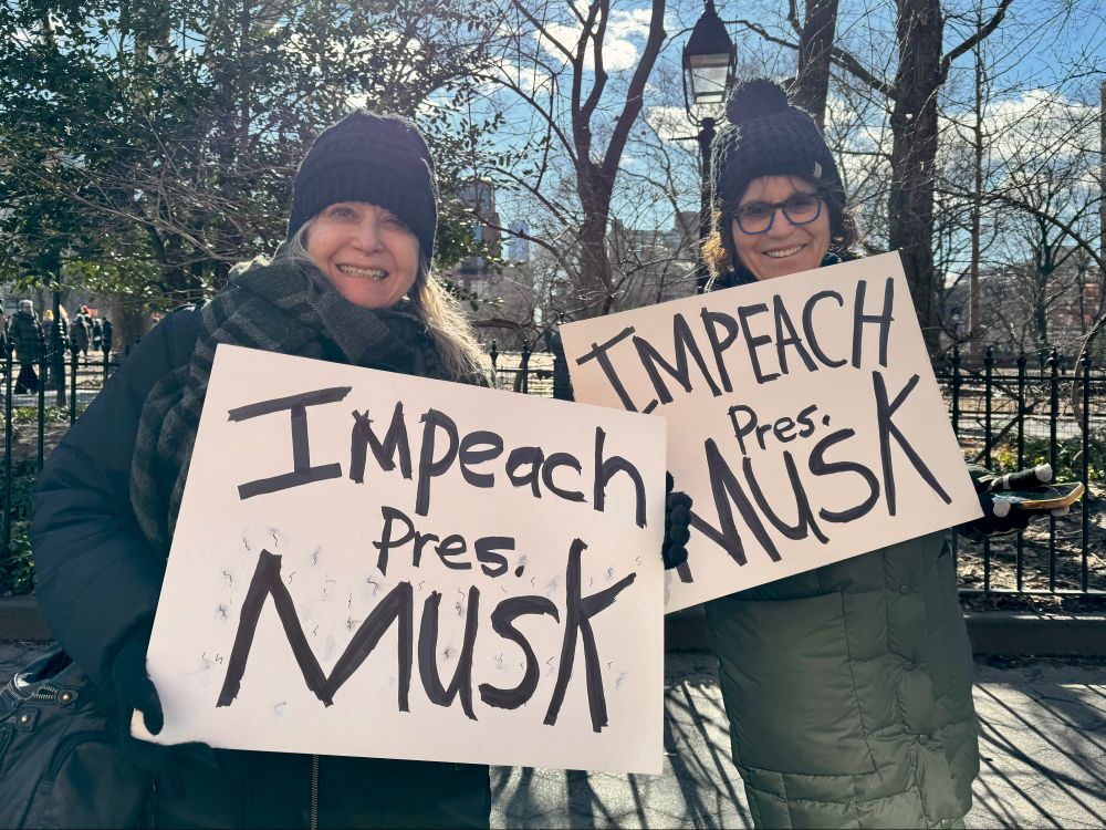 two women hold up handmade cardboard signs that say IMPEACH PRES. MUSK